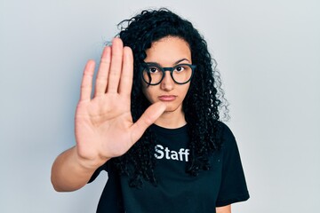 Young hispanic woman with curly hair wearing staff t shirt doing stop sing with palm of the hand....