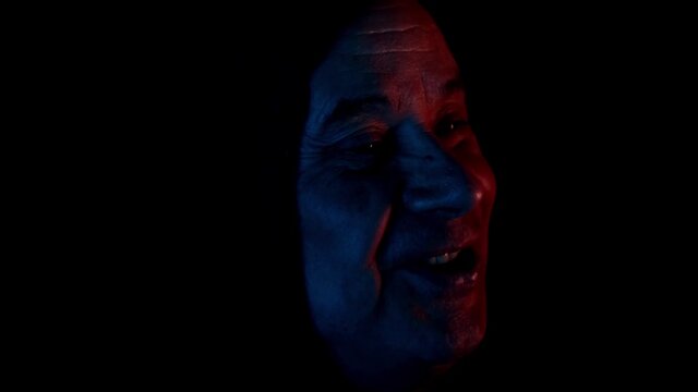 elderly man in a dark room with neon lights in a black hood sits, watches TV, grins, head close-up, concept mood, modern technologies, emotions, addiction, adrenaline