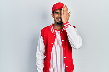 Young african american man wearing baseball uniform covering one eye with hand, confident smile on face and surprise emotion.