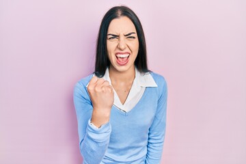 Beautiful woman with blue eyes standing over pink background angry and mad raising fist frustrated...