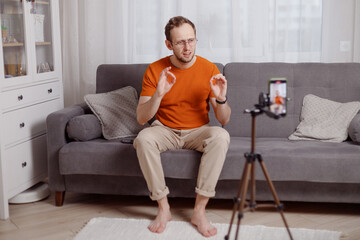 Fototapeta premium Hipster man blogger recording vlog on mobile phone sit on couch in living room and explain and gesturing with hands