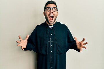 Young hispanic man wearing priest uniform standing over white background crazy and mad shouting and...