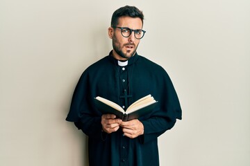 Young hispanic priest man holding bible clueless and confused expression. doubt concept.
