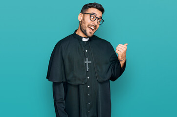 Young hispanic man wearing priest uniform smiling with happy face looking and pointing to the side...