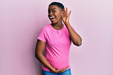 Young african american woman expecting a baby, touching pregnant belly smiling with hand over ear listening an hearing to rumor or gossip. deafness concept.