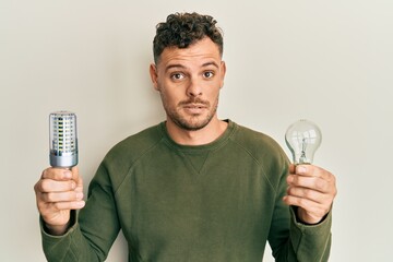 Young hispanic man holding lightbulb for inspiration and idea clueless and confused expression....