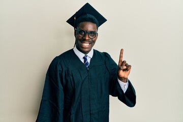 Handsome black man wearing graduation cap and ceremony robe pointing finger up with successful...