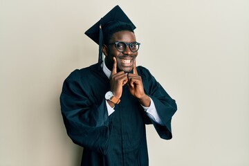 Handsome black man wearing graduation cap and ceremony robe smiling with open mouth, fingers...