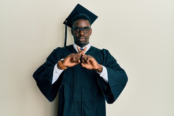 Handsome black man wearing graduation cap and ceremony robe rejection expression crossing fingers doing negative sign