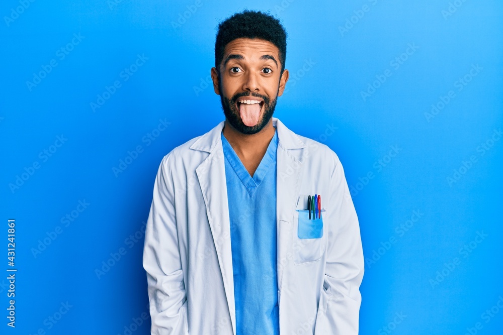 Wall mural Handsome hispanic man with beard wearing doctor uniform sticking tongue out happy with funny expression. emotion concept.