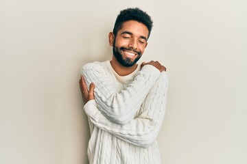 Handsome hispanic man with beard wearing casual winter sweater hugging oneself happy and positive,...