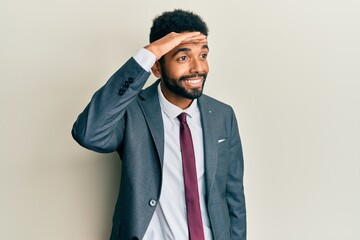 Handsome hispanic man with beard wearing business suit and tie very happy and smiling looking far away with hand over head. searching concept.