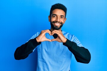 Handsome hispanic man with beard wearing blue male nurse uniform smiling in love showing heart symbol and shape with hands. romantic concept.