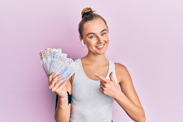 Beautiful blonde sport woman holding 20 swedish krona banknotes smiling happy pointing with hand and finger