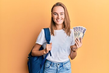 Beautiful blonde woman wearing student backpack and holding 5000 japan yen looking positive and happy standing and smiling with a confident smile showing teeth