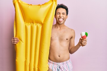 Obraz premium Young handsome man wearing swimsuit holding summer mattress float and icecream sticking tongue out happy with funny expression.