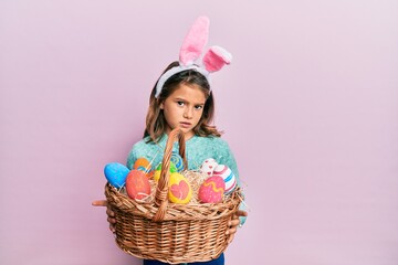 Little beautiful girl wearing cute easter bunny ears holding wicker basket with colored eggs clueless and confused expression. doubt concept.