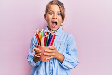 Little beautiful girl holding colored pencils celebrating crazy and amazed for success with open...