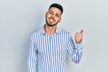 Naklejka premium Young hispanic man with beard wearing casual striped shirt smiling with happy face looking and pointing to the side with thumb up.