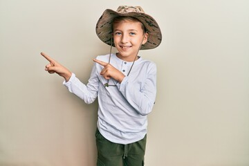 Little caucasian boy kid wearing explorer hat smiling and looking at the camera pointing with two hands and fingers to the side.