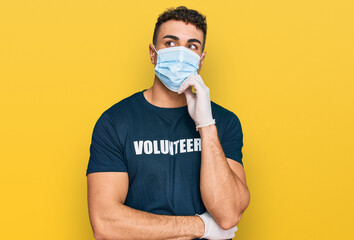 Hispanic young man wearing medical mask and volunteer t shirt serious face thinking about question...