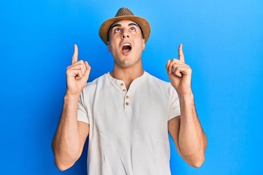 Hispanic young man wearing summer hat amazed and surprised looking up and pointing with fingers and raised arms.