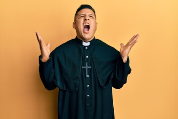 Young latin priest man standing over yellow background crazy and mad shouting and yelling with...