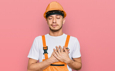 Hispanic young man wearing handyman uniform and safety hardhat smiling with hands on chest with closed eyes and grateful gesture on face. health concept.