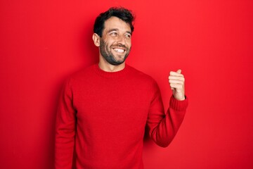 Fototapeta premium Handsome man with beard wearing casual red sweater smiling with happy face looking and pointing to the side with thumb up.