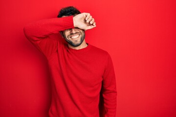 Handsome man with beard wearing casual red sweater covering eyes with arm smiling cheerful and funny. blind concept.