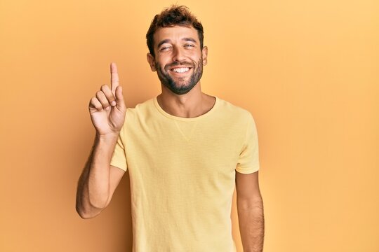 Handsome man with beard wearing casual yellow tshirt over yellow background showing and pointing up with finger number one while smiling confident and happy.