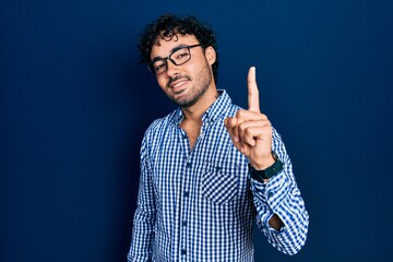Young hispanic man wearing casual clothes and glasses smiling with an idea or question pointing...