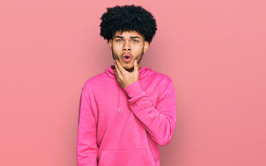 Obraz na płótnie Canvas Young african american man with afro hair wearing casual pink sweatshirt looking fascinated with disbelief, surprise and amazed expression with hands on chin