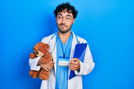 Young hispanic man wearing doctor uniform holding teddy bear and clipboard smiling looking to the side and staring away thinking.