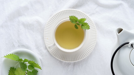 Nettle tea cup, fresh stinging nettle herbs. Flat lay, top view, off white textile tablecloth. White ceramic teapot. Natural light.