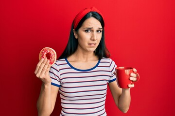 Young hispanic woman eating doughnut and drinking coffee clueless and confused expression. doubt...