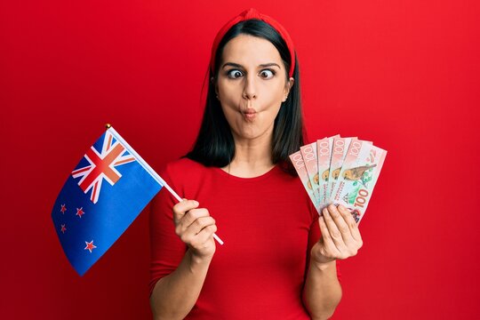 Young hispanic woman holding new zealand flag and dollars making fish face with mouth and squinting eyes, crazy and comical.