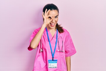 Young brunette woman wearing doctor uniform and stethoscope doing ok gesture with hand smiling, eye looking through fingers with happy face.