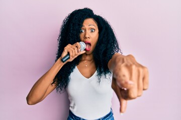 Middle age african american woman singing song using microphone pointing with finger afraid and...