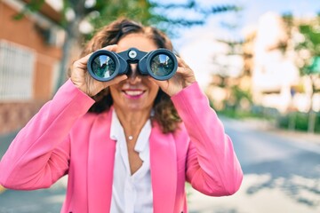 Middle age hispanic businesswoman smiling happy looking for new opportunity using binoculars at the city.