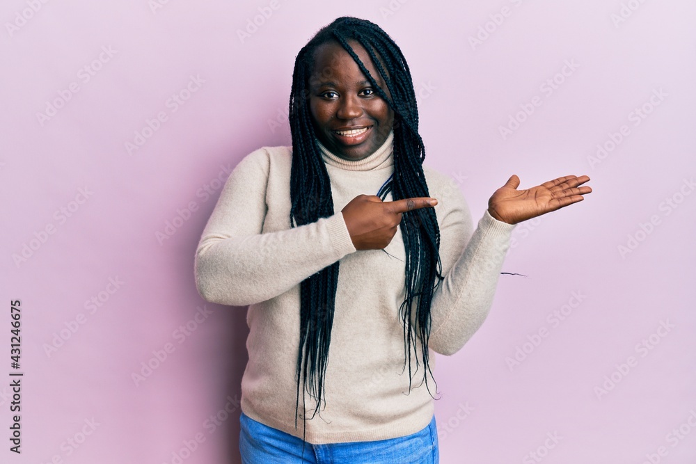 Sticker Young black woman with braids wearing casual winter sweater amazed and smiling to the camera while presenting with hand and pointing with finger.