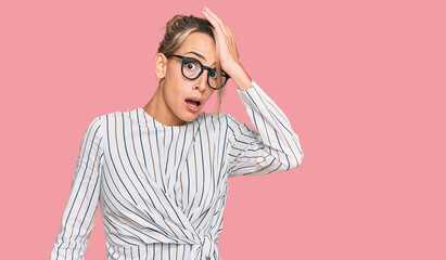 Beautiful blonde woman wearing business shirt and glasses surprised with hand on head for mistake,...