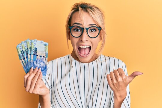 Beautiful blonde woman holding south african 100 rand banknotes pointing thumb up to the side smiling happy with open mouth