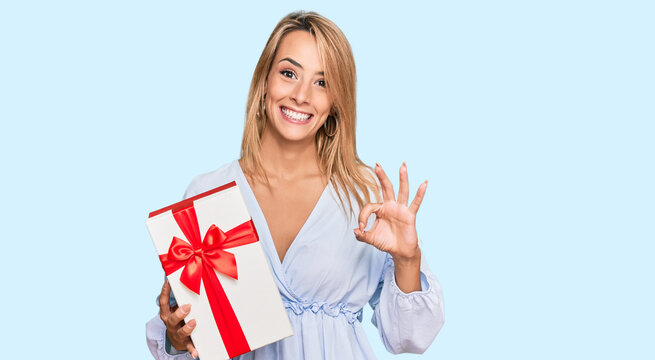 Beautiful blonde woman holding gift doing ok sign with fingers, smiling friendly gesturing excellent symbol
