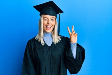 Beautiful blonde woman wearing graduation cap and ceremony robe smiling with happy face winking at the camera doing victory sign. number two.