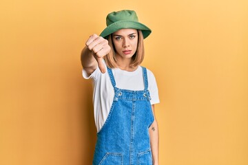 Young caucasian blonde woman wearing denim jumpsuit and hat with 90s style looking unhappy and...