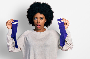 Young african american woman holding socks in shock face, looking skeptical and sarcastic, surprised with open mouth