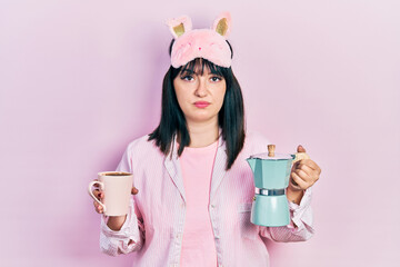 Young hispanic woman wearing sleep mask and robe drinking coffee relaxed with serious expression on...