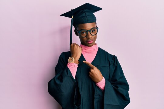Young african american girl wearing graduation cap and ceremony robe in hurry pointing to watch time, impatience, looking at the camera with relaxed expression