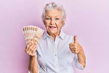 Senior grey-haired woman holding 50 turkish lira banknotes smiling happy and positive, thumb up...